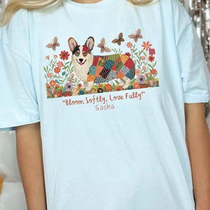Może przedstawiać: Jasnoniebieska koszulka z kolorową ilustracją psa corgi z patchworkowym wzorem. Projekt zawiera kwiaty, motyle oraz tekst "Bloom Softly, Love Fully" i "Sasha".