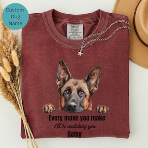 May include: Burgundy-colored t-shirt featuring a German Shepherd dog illustration. The shirt has the text "Every move you make I'll be watching you Daisy". A silver necklace with a star pendant is draped on the shirt. A custom dog name can be added.