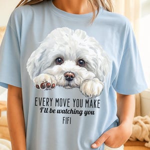 May include: Light blue t-shirt featuring a white, fluffy dog illustration with brown eyes and paws. The text "EVERY MOVE YOU MAKE I'll be watching you FIFI" is printed below the dog.