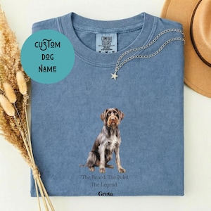 May include: A blue crew neck t-shirt featuring a graphic of a brown and white German Wirehaired Pointer dog. Below the dog, the text reads, "The Beard. The Point. The Legend." A silver star-shaped pendant necklace rests on the collar area.