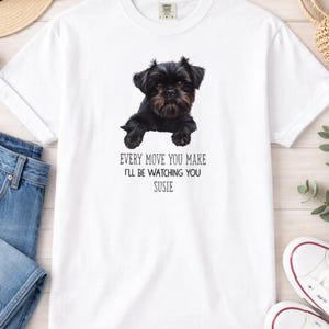 May include: White t-shirt featuring a black and brown dog illustration. The text below the dog reads, "EVERY MOVE YOU MAKE I'LL BE WATCHING YOU SUSIE." The shirt is made of a soft, comfortable material.