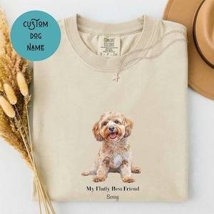 Könnte beinhalten: Ein beiges T-Shirt mit Rundhalsausschnitt und einer Aquarell-Illustration eines sitzenden hellbraunen Hundes. Unter dem Hund steht der Text "My Fluffy Best Friend" und "Benny". Das T-Shirt hat ein Comfort Colors Etikett am Innenkragen.