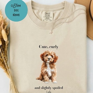 Op de afbeelding: Een beige t-shirt met ronde hals en een afbeelding van een bruine en witte puppy met krullend haar. De tekst boven de puppy luidt "Cute, curly" en de tekst eronder luidt "and slightly spoiled" gevolgd door de naam "Lulu" in een cursief lettertype.