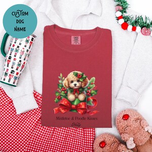 Op de afbeelding: Een rood t-shirt met een afbeelding van een poedelpuppy in een groene bak omringd door hulst en een rode strik, met de tekst Mistletoe & Poodle Kisses en de naam Holly onder de illustratie op de voorzijde van het kledingstuk.