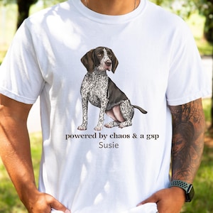 Pode incluir: Camiseta branca com uma ilustração de um cão de raça Braco Alemão sentado, com marcas castanhas e brancas. O texto "powered by chaos & a gsp Susie" está impresso abaixo.