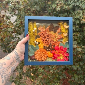 Op de afbeelding: Een blauw omkaderde herfst-thema schaduwdoos met de tekst "Happy Fall". De doos bevat oranje en rode bloemen, vlinders en herfstbladeren. Een kleine pompoen en decoratief mos maken de herfst esthetiek compleet.