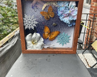 May include: A framed shadow box with a brown wooden frame. Inside, there are two orange butterflies, blue and white flowers, a silver snowflake, and a white flower. The background is a dark blue with glitter.