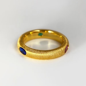May include: A gold ring with a textured surface, featuring a blue, green, and red gemstone. The ring is set against a white background, highlighting its vibrant colours and design.
