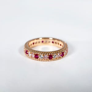 Peut inclure: Une bague en or rose ornée d'une rangée continue de pierres précieuses rouges, roses et claires alternées. La bague présente un motif de corde tressée sur les bords, sur fond blanc.