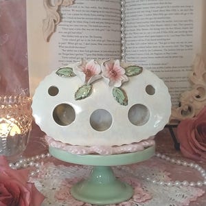 May include: A cream-coloured ceramic holder with several circular openings, decorated with pink floral details and green leaves. It sits on a pale green pedestal base with a pink lace trim. The holder is designed for decorative purposes.