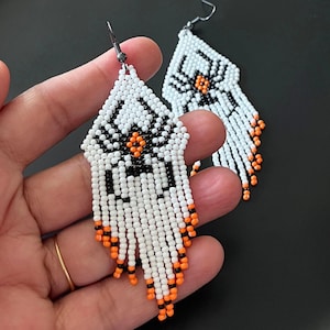 Puede incluir: Pendientes de cuentas hechos a mano con un diseño de araña. Los pendientes son blancos con detalles en negro y naranja. La araña está en el centro de los pendientes. La borla en la parte inferior es blanca con cuentas naranjas y negras.