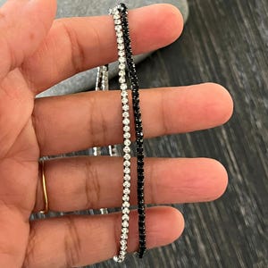 May include: Two delicate tennis bracelets, one silver-toned with clear crystals and the other black with black crystals. The bracelets are held up against a dark background, showcasing their sparkle and design. A gold ring is visible on the hand.