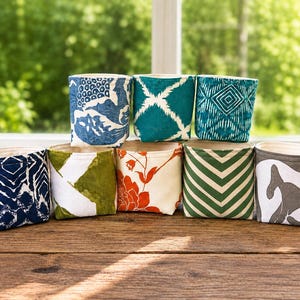 Puede incluir: Una colección de ocho cestas de almacenamiento cilíndricas de tela con varios patrones y colores, incluyendo diseños florales en azul y blanco, geométricos en turquesa, florales en naranja, chevron en verde y abstractos en gris, en dos filas.