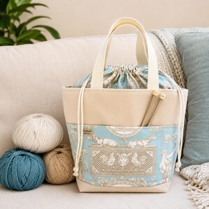 May include: A beige and light blue fabric project bag with a drawstring closure and two top handles. The lower section features a repeating toile-style pattern of dogs and classical motifs. Two wooden knitting needles are tucked into the front pocket.
