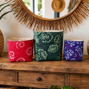 May include: Three fabric storage baskets with a cylindrical shape and a light-colored interior lining. The left basket is red with a white spiral pattern, the center basket is dark green with a light green leaf print, and the right basket is purple with a blue floral pattern.