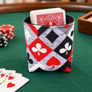 May include: A fabric card holder featuring a repeating diamond pattern in red, black, and grey, decorated with white and black playing card suit symbols including clubs, spades, and hearts, containing a deck of playing cards.