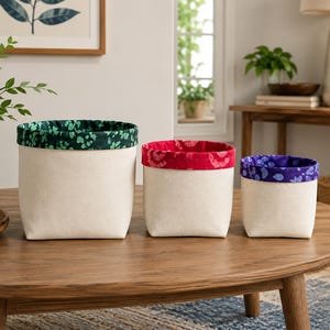 May include: Three off-white fabric storage bins with patterned trim in green, red, and blue. The bins are cylindrical with a square base and sit on a wooden table. The trim features floral or abstract designs.