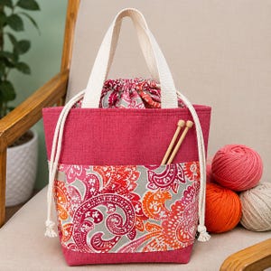 May include: A magenta fabric tote bag with a drawstring closure and a front pocket featuring a paisley pattern in orange, red, and gray. The bag has two cream-colored woven handles and two wooden knitting needles tucked into the front pocket.