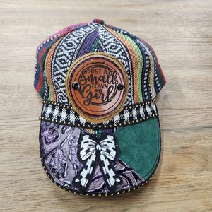 May include: A baseball cap with a colorful patterned top, a brown leather patch with the text "Just a Small Town Girl", and a decorative bow. The brim features a green suede patch and a purple patterned section.