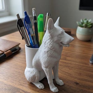 May include: A white, three-dimensional desk organizer shaped like a sitting German Shepherd dog. The cylindrical container portion holds several pens, pencils, and a highlighter. The dog figure is sculpted with detailed fur texture and pointed ears.