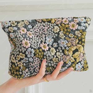 以下が含まれることがあります： 黒い背景に花柄のクラッチバッグ。ピンク、黄色、青の色合いの刺繍の花が施されたキルティングデザインが特徴です。バッグは手で持たれており、生地の複雑なディテールが示されています。