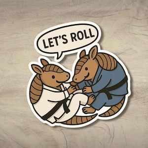 May include: A sticker depicting two cartoon armadillos in martial arts attire. One wears a white gi with a black belt, the other a blue gi with a black belt. A speech bubble above the armadillos reads "LET'S ROLL".
