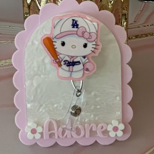 May include: A pink badge reel with a scalloped edge and a white and pink marbled background. The reel features a Hello Kitty character in a Dodgers baseball uniform, holding a bat. The word "Adore" is written in pink.