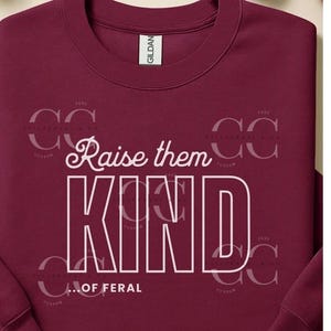 May include: Burgundy crewneck sweatshirt with the text "Raise them KIND...OF FERAL" in white block letters. The sweatshirt has a Gildan tag and a subtle design in the background. A casual, comfortable top.