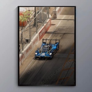 May include: A framed print featuring a blue race car with the number 23, speeding along a track. The car has the Aston Martin logo. The track is bordered by a red and white barrier with the words "TIRE RACK". The image is labeled "SUPERCARSHOTS".