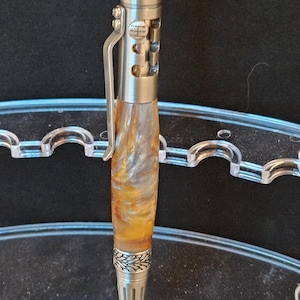 Puede incluir: Un bolígrafo artesanal con un cuerpo de metal plateado y un barril que presenta una mezcla de tonos naranja, amarillo y blanco. El bolígrafo tiene un clip y detalles decorativos, incluyendo una parte superior e inferior estampadas. El bolígrafo se exhibe sobre un soporte transparente y curvado.