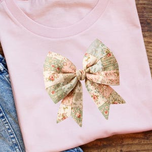 May include: A pale pink t-shirt with a large bow applique. The bow is made of fabric patches in pink, green, and cream, with a floral pattern. The shirt is displayed on a wooden surface.