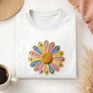 May include: White t-shirt with a large, colorful daisy applique. The daisy features yellow, pink, and blue petals with a textured, knitted appearance. The center of the daisy is a spiral of yellow yarn. A cup of coffee and a straw hat are in the background.