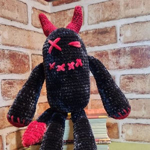 May include: A handmade black crochet monster doll with red accents. The doll has horns, crossed-out eyes, and a stitched mouth. The arms and legs are attached, and the feet are red. The doll is sitting on a stack of books.