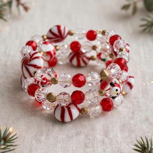 May include: A multi-strand coil bracelet featuring red and white peppermint-style beads, clear faceted crystal beads, small metallic gold spacer beads, and a small snowman-shaped bead with a black hat and orange nose detail.