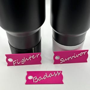 May include: Three pink tags with white lettering that read "Fighter", "Survivor", and "Badass". The tags are in front of two black tumblers.