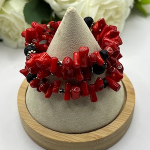 May include: A multi-strand wrap bracelet featuring irregular, bright red coral-like beads interspersed with porous black lava stone beads and small metallic silver spacers, arranged in a layered design on a conical display stand.