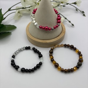 Pode incluir: Três pulseiras de contas. Uma apresenta contas prateadas e fúcsia, outra contas pretas e brancas, e a terceira contas castanhas e douradas. As pulseiras são exibidas numa superfície branca. As contas são medidas em milímetros.