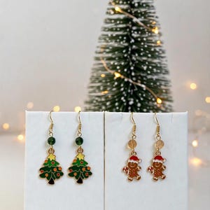 May include: Two pairs of dangle earrings on white display cards. One pair features green Christmas tree charms with yellow stars and red ornaments. The other pair features brown gingerbread man charms wearing red Santa hats. Both pairs have gold-tone hooks.