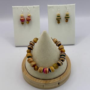 May include: A jewelry set consisting of a beaded bracelet and two pairs of drop earrings. The bracelet features round beads in natural wood, tiger eye, and multi-colored striped patterns. The earrings match, with one pair featuring pink stripes and the other green stripes, all suspended from silver-toned metal hooks.