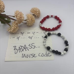 Puede incluir: Dos pulseras de cuentas y una nota con texto escrito a mano. Una pulsera presenta cuentas rojas y blancas translúcidas con un acento plateado. La otra tiene cuentas negras y blancas veteadas. La nota dice: ... - .-. ... -.. .- ... ... Morse code.