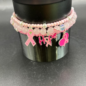 May include: A multi-strand beaded bracelet featuring light pink seed beads, white pearls, and translucent pink stone beads. The bracelet includes three metal charms: a pink ribbon with the word WARRIOR, the word HOPE, and a pink heart ribbon.