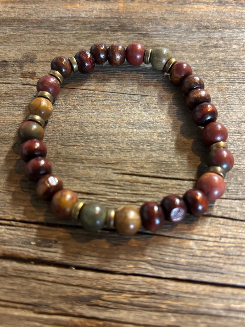 Puede incluir: Una pulsera el&aacute;stica compuesta por cuentas redondas en tonos marr&oacute;n oscuro, marr&oacute;n rojizo y verde oliva apagado. Las cuentas est&aacute;n intercaladas con peque&ntilde;os espaciadores met&aacute;licos planos de color lat&oacute;n que crean un patr&oacute;n circular repetitivo.
