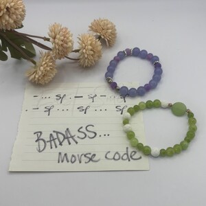May include: Two beaded bracelets and a note. One bracelet features purple beads with silver accents. The other has green beads and white textured beads. The note displays Morse code and the handwritten text "BADASS... Morse code".