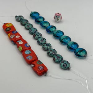 May include: A collection of beads including a strand of rectangular orange beads with floral patterns, a strand of round teal beads with floral designs, a strand of round blue glass beads, and a single small white and pink owl-shaped bead.