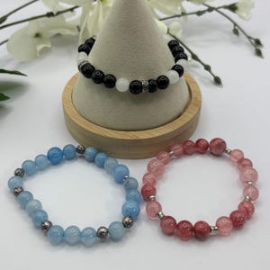 May include: Three beaded bracelets. One bracelet features black and white beads with silver accents, displayed on a wooden stand. The other two bracelets are light blue and pink, respectively, with silver accents. The bracelets are on a white surface.