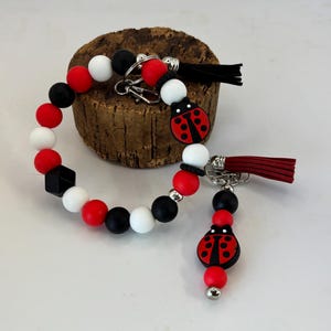 Op de afbeelding: Een kralen sleutelhanger en armband set met rode, zwarte en witte kralen. De sleutelhanger heeft een lieveheersbeestje bedel en een rode kwast. De armband heeft een zwarte vierkante kraal. Beide hebben zilverkleurige hardware.