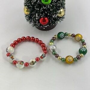 May include: Two beaded bracelets positioned below a small artificial Christmas tree. The left bracelet features red spherical beads and white beads with nutcracker illustrations. The right bracelet consists of green, gold, and silver beads with white snowflake patterns.