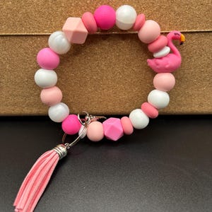 May include: A pink beaded keychain wristlet with a flamingo charm. The wristlet features a variety of pink and white beads, including round and geometric shapes. A pink tassel hangs from the keychain.