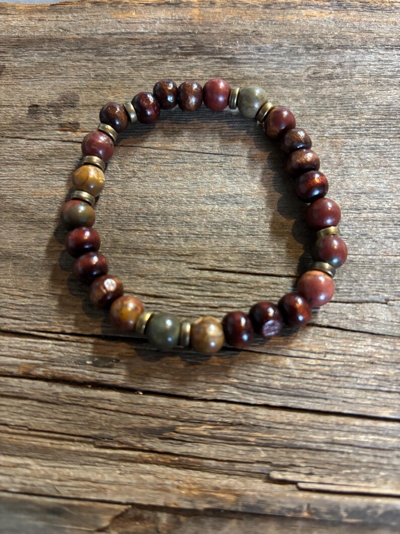 Puede incluir: Una pulsera el&aacute;stica compuesta por cuentas redondas y pulidas en tonos rojo intenso, marr&oacute;n y verde apagado, intercaladas con peque&ntilde;os espaciadores met&aacute;licos cil&iacute;ndricos. Las cuentas var&iacute;an en intensidad de color y textura.