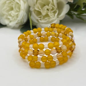 May include: A multi-strand wrap bracelet featuring a combination of matte yellow beads, translucent white beads, and faceted amber-colored square beads. The design includes small metallic gold textured spherical accents throughout the spiral structure.